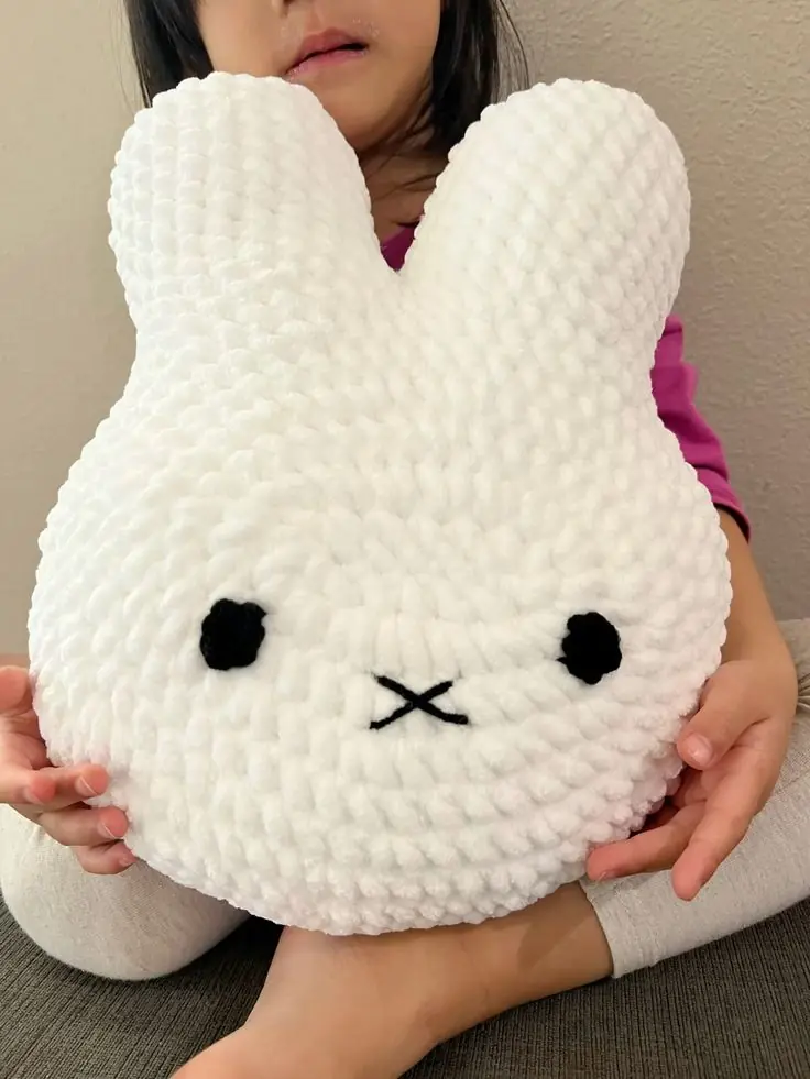 A large white crochet bunny-face pillow with black eyes and a cross-stitched mouth