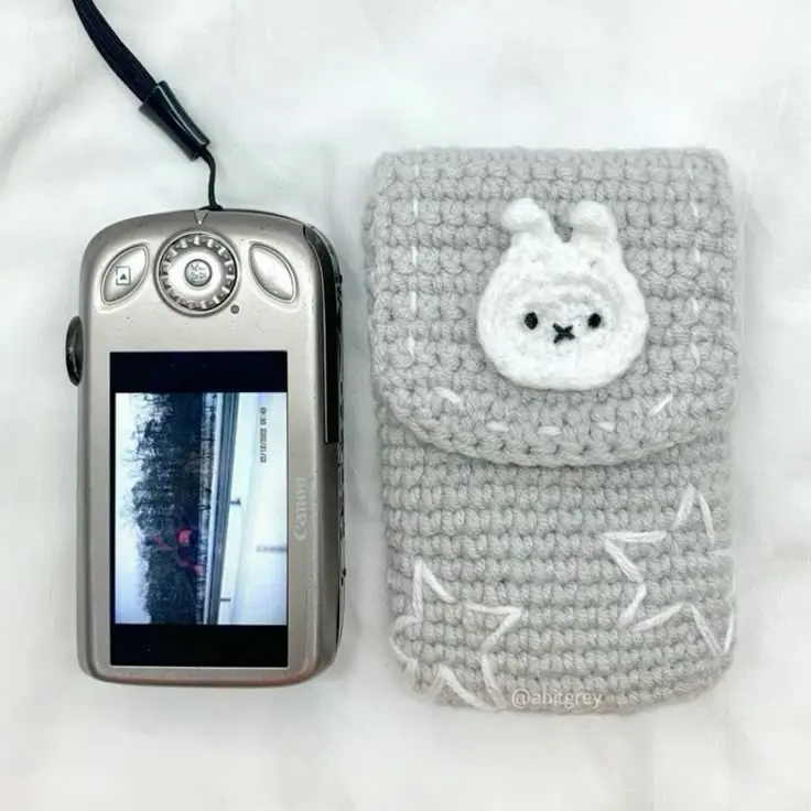 A grey crochet camera pouch with a small bunny face appliqué, next to a digital camera