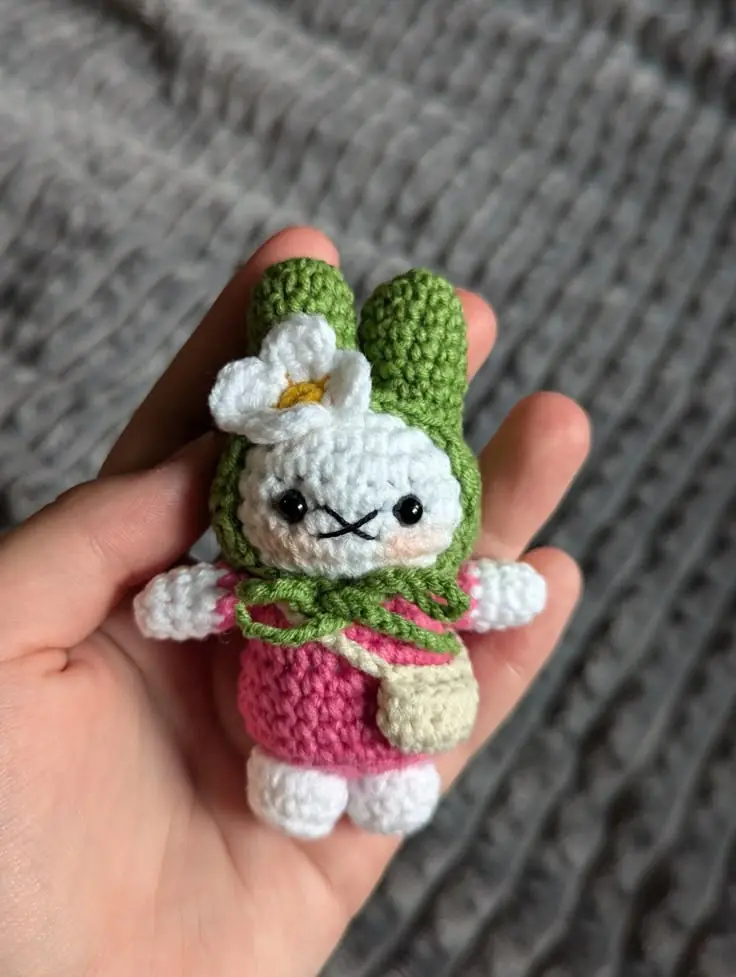 A tiny crochet bunny in a green hood and pink dress, with a white flower, held in a hand