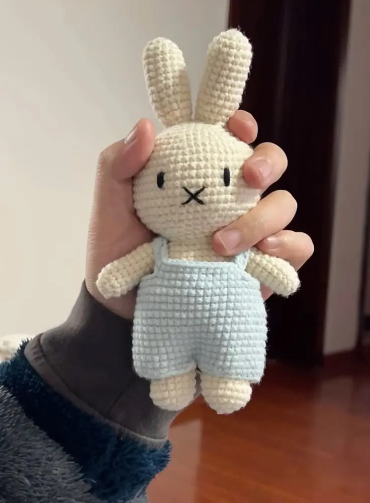 A cream crochet bunny wearing light blue overalls, held in a hand