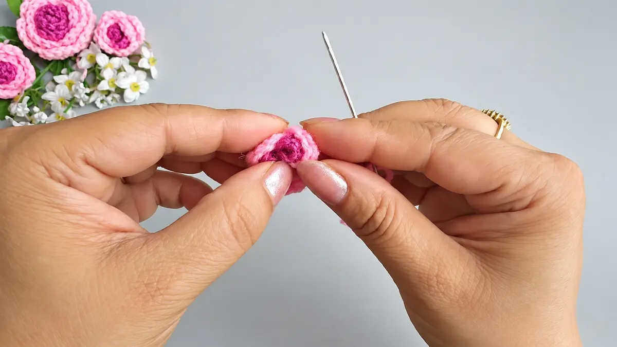Hands shaping a small pink crochet rose with a yarn needle